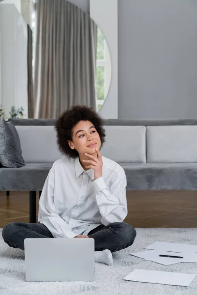 Verträumte afrikanisch-amerikanische Frau sitzt neben Laptop und Dokumenten auf Teppich neben grauem Samtsofa — Stockfoto