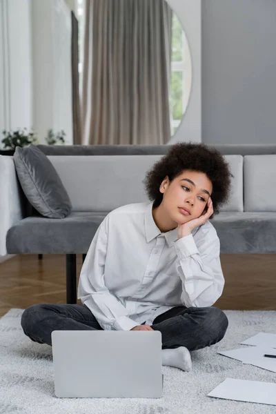 Gelangweilte Afroamerikanerin sitzt neben Laptop und Dokumenten auf Teppich neben grauem Samtsofa — Stockfoto