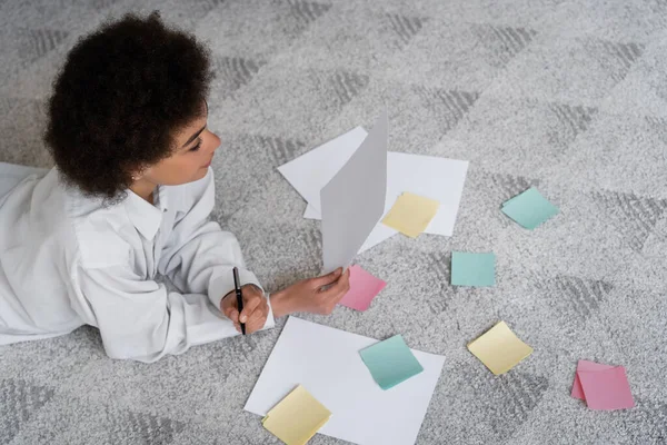 Vista superior de la mujer afroamericana rizada sosteniendo documento cerca de papeles en blanco en la alfombra - foto de stock