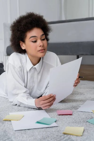 Curly african american woman holding blank document near papers on carpet — Stock Photo