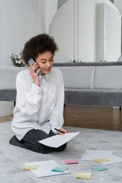 Mujer afroamericana hablando en teléfono inteligente mientras mira el documento cerca de papeles en blanco en la alfombra — Stock Photo