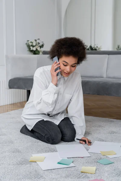 Кудрявая африканская американка разговаривает на смартфоне, глядя на документ рядом с пустыми бумагами на ковре — стоковое фото
