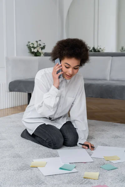 Lockige afrikanisch-amerikanische Frau spricht auf dem Smartphone, während sie Dokument in der Nähe leere Papiere auf dem Teppich betrachtet — Stockfoto