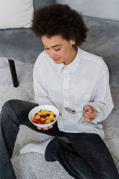 Vista ad alto angolo della donna afro-americana che tiene ciotola con fiocchi di mais e bacche — Foto stock