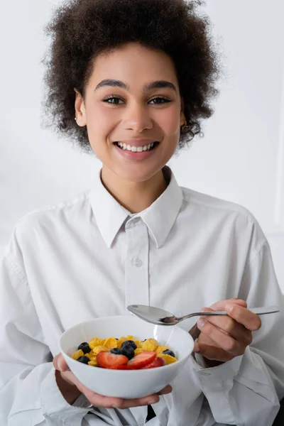 Cheerful african american woman holding bowl with corn flakes and berries — Stock Photo