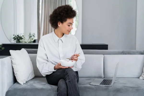African american woman holding bowl with breakfast while looking at laptop on velvet sofa — Stock Photo
