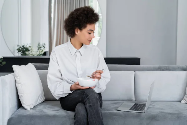 Afrikanerin hält Schüssel mit Frühstück in der Hand und blickt auf Laptop auf Samtsofa — Stockfoto