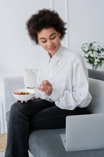 Pleased african american woman holding bowl with berries while looking at laptop on velvet sofa — Stock Photo