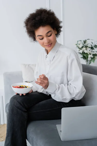 Mulher afro-americana satisfeito segurando tigela com bagas enquanto olha para laptop no sofá de veludo — Fotografia de Stock