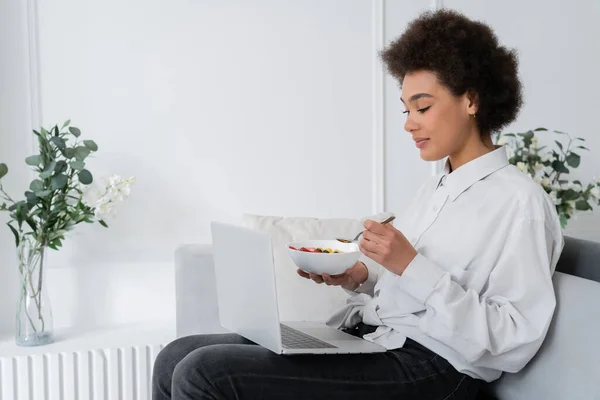 Donna afroamericana che tiene la ciotola con le bacche mentre guarda il film sul computer portatile e sul divano di velluto seduto — Foto stock