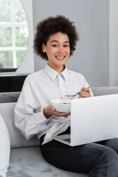 Heureuse femme afro-américaine tenant bol avec petit déjeuner tout en regardant la caméra et assis avec ordinateur portable sur canapé en velours — Photo de stock
