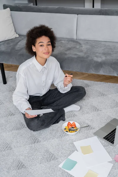 Curly african american freelancer holding pen and paper while sitting on carpet near bowl with breakfast and laptop — Stock Photo