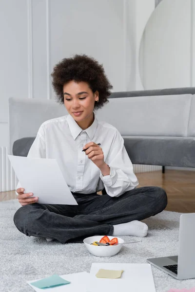 Young african american freelancer holding pen and paper while sitting on carpet near bowl with breakfast and laptop — Stock Photo