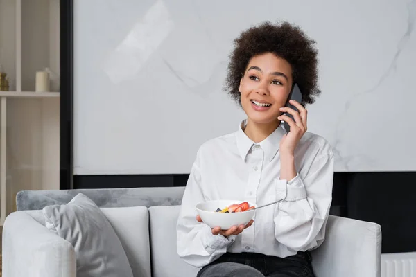 Felice donna afroamericana che tiene la ciotola con la colazione e parla sullo smartphone — Foto stock