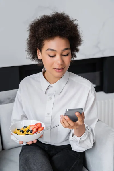 Femme afro-américaine bouclée tenant bol avec petit déjeuner et smartphone — Photo de stock