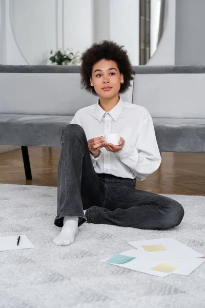 Кучерява афроамериканка тримає чашку кави і сидить біля документів на килимі — стокове фото