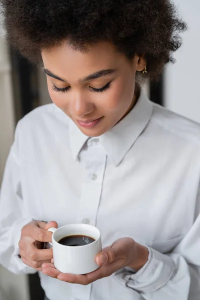 Ricci donna afroamericana in camicia bianca guardando una tazza di caffè — Foto stock