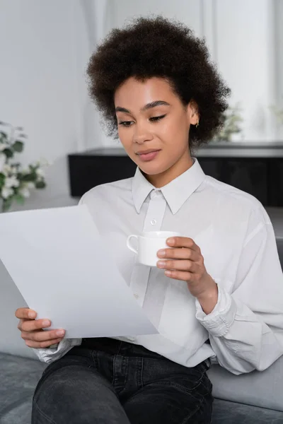 Mujer afroamericana rizada sosteniendo una taza de café mientras mira el papel en blanco - foto de stock