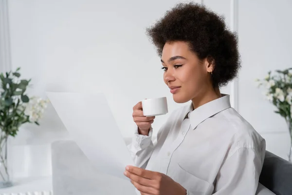 Encaracolado afro-americano mulher segurando xícara de café enquanto olha para o documento em branco — Fotografia de Stock