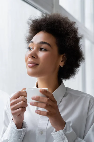 Mujer afroamericana rizada y sonriente sosteniendo una taza de café - foto de stock