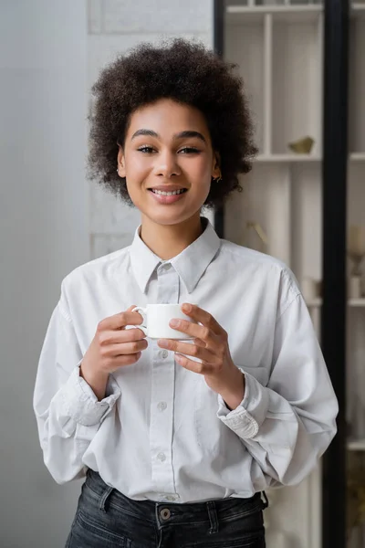 Freudige und lockige afrikanisch-amerikanische Frau, die eine Tasse Kaffee hält und in die Kamera schaut — Stockfoto