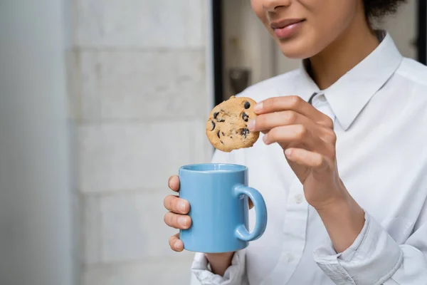 Vue recadrée d'une femme afro-américaine heureuse tenant une tasse de lait et un cookie avec des pépites de chocolat — Photo de stock