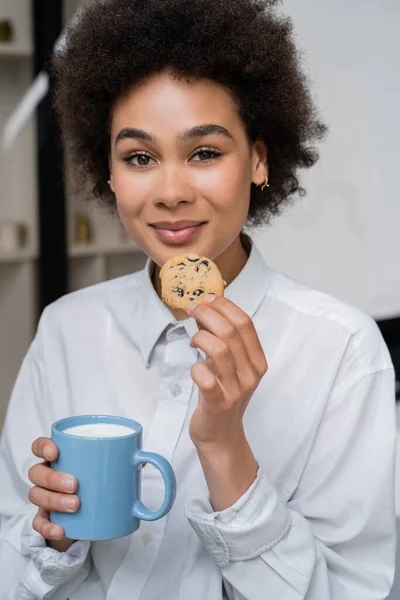 Alegre mujer afroamericana sosteniendo taza de leche y galletas con chispas de chocolate - foto de stock