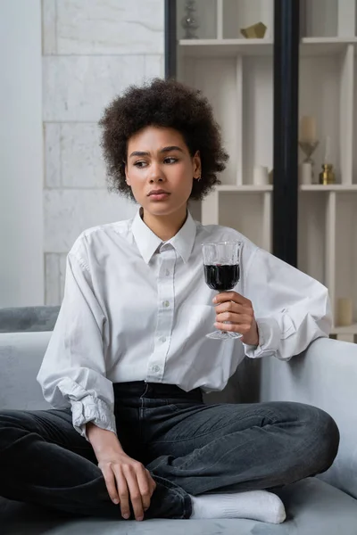 Sad young african american woman holding glass of red wine while sitting on velvet sofa — Stock Photo