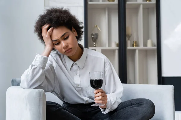 Molesto joven afroamericana mujer sosteniendo vaso de vino tinto mientras está sentado en un sofá de terciopelo - foto de stock