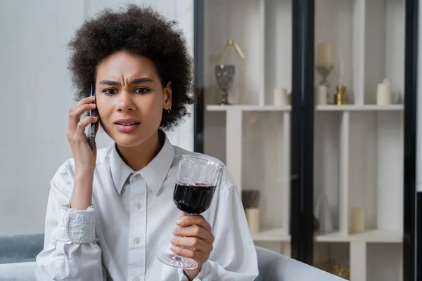 Triste donna afroamericana che tiene in mano un bicchiere di vino rosso e parla sullo smartphone — Foto stock