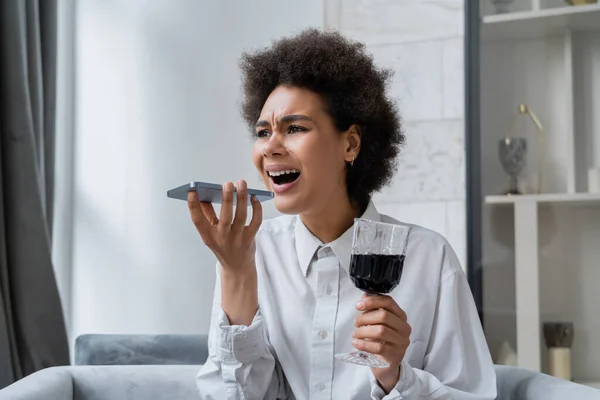 Ivre et triste femme afro-américaine tenant un verre de vin rouge tout en enregistrant un message vocal sur smartphone — Photo de stock