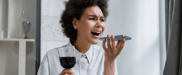 Betrunkene und traurige Afroamerikanerin hält ein Glas Rotwein in der Hand, während sie eine Sprachnachricht auf ihrem Smartphone aufzeichnet, Banner — Stockfoto