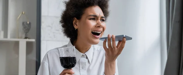 Drunk and sad african american woman holding glass of red wine while recording voice message on smartphone, banner — Stock Photo
