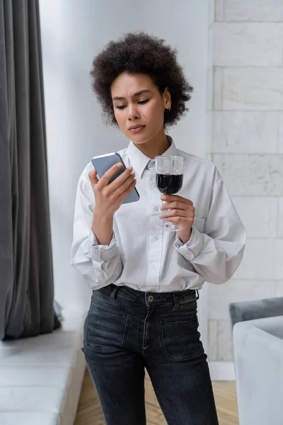 Triste afro-americana segurando copo de vinho tinto e olhando para smartphone — Fotografia de Stock