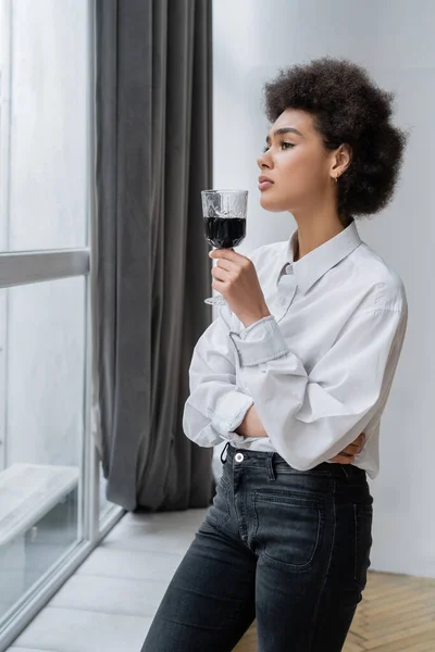Triste femme afro-américaine tenant un verre de vin rouge et regardant la fenêtre — Photo de stock
