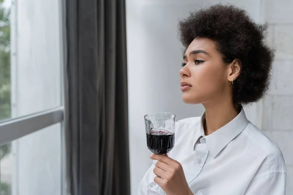 Mujer afroamericana molesta sosteniendo un vaso de vino tinto y mirando a la ventana - foto de stock
