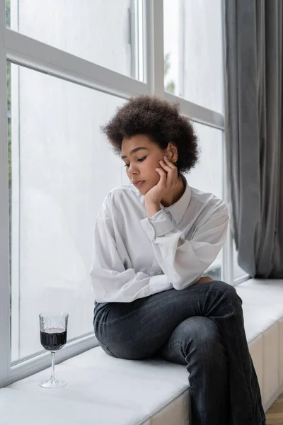 Mujer afroamericana triste mirando un vaso de vino tinto mientras está sentada en el alféizar de la ventana - foto de stock