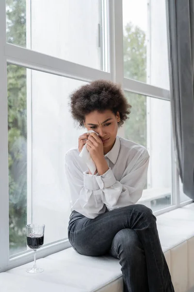 Triste femme afro-américaine essuyant des larmes près du verre de vin rouge tout en étant assis sur le rebord de la fenêtre — Photo de stock