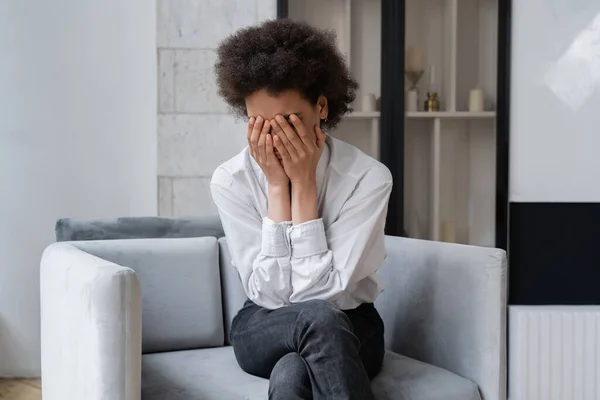 Stressato donna afroamericana in camicia bianca che copre il viso mentre seduto in soggiorno — Foto stock