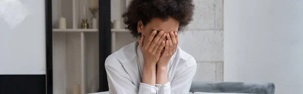 Stressed african american woman in white shirt covering face in living room, banner — Stock Photo