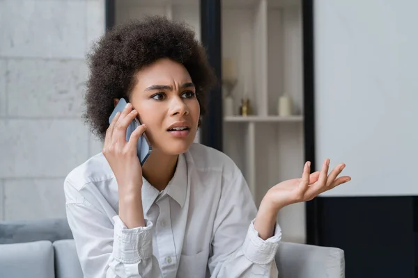 Mujer afroamericana disgustada en camisa blanca hablando en smartphone y haciendo gestos en casa — Stock Photo