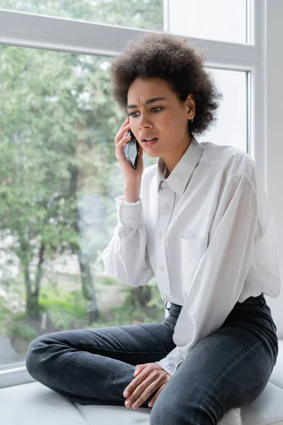 Donna afro-americana scontenta in camicia bianca che parla sullo smartphone vicino alla finestra — Stock Photo