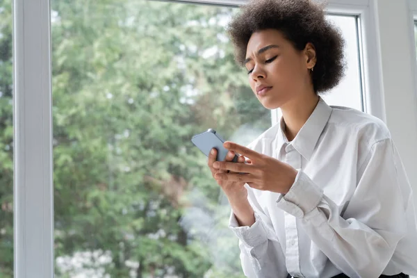 Vista de ángulo bajo de la mujer afroamericana molesta en camisa blanca mensaje de lectura en el teléfono inteligente cerca de la ventana - foto de stock