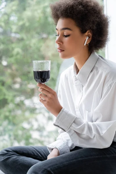 Triste femme afro-américaine en chemise blanche et écouteur sans fil regardant un verre de vin rouge — Photo de stock