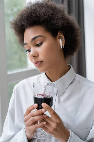 Femme américaine africaine déprimée en chemise blanche et écouteur sans fil tenant un verre de vin rouge — Photo de stock