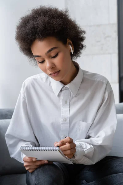 Curly african american woman in white shirt and wireless earphone writing in notebook — Stock Photo