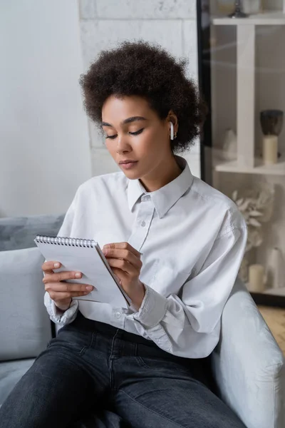 Femme afro-américaine bouclée en chemise blanche et écouteur sans fil prenant des notes dans le carnet — Photo de stock