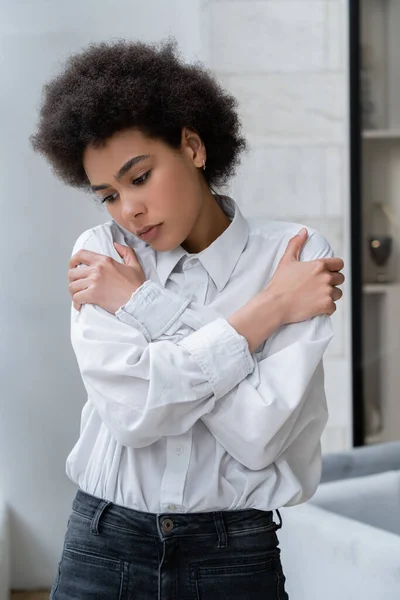 Depressed african american woman in white shirt hugging shoulders at home — Stock Photo