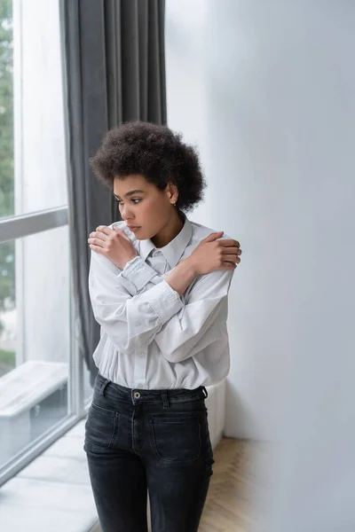 Depressed african american woman in white shirt and jeans hugging shoulders at home — Stock Photo