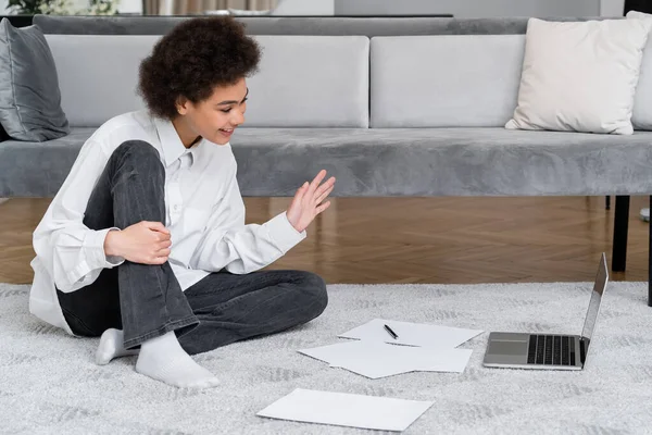 Feliz afroamericana mujer teniendo video chat en el ordenador portátil mientras está sentado cerca de terciopelo gris sofá - foto de stock